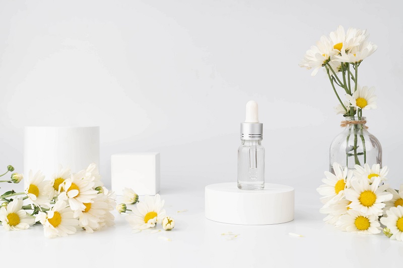 Mock up produs cosmetic natural pentru îngrijirea pielii. Flacon cosmetic de sticlă cu picurător și floare organică de primăvară pe podium cu cerc alb. Recipient de ser, esență sau parfum.